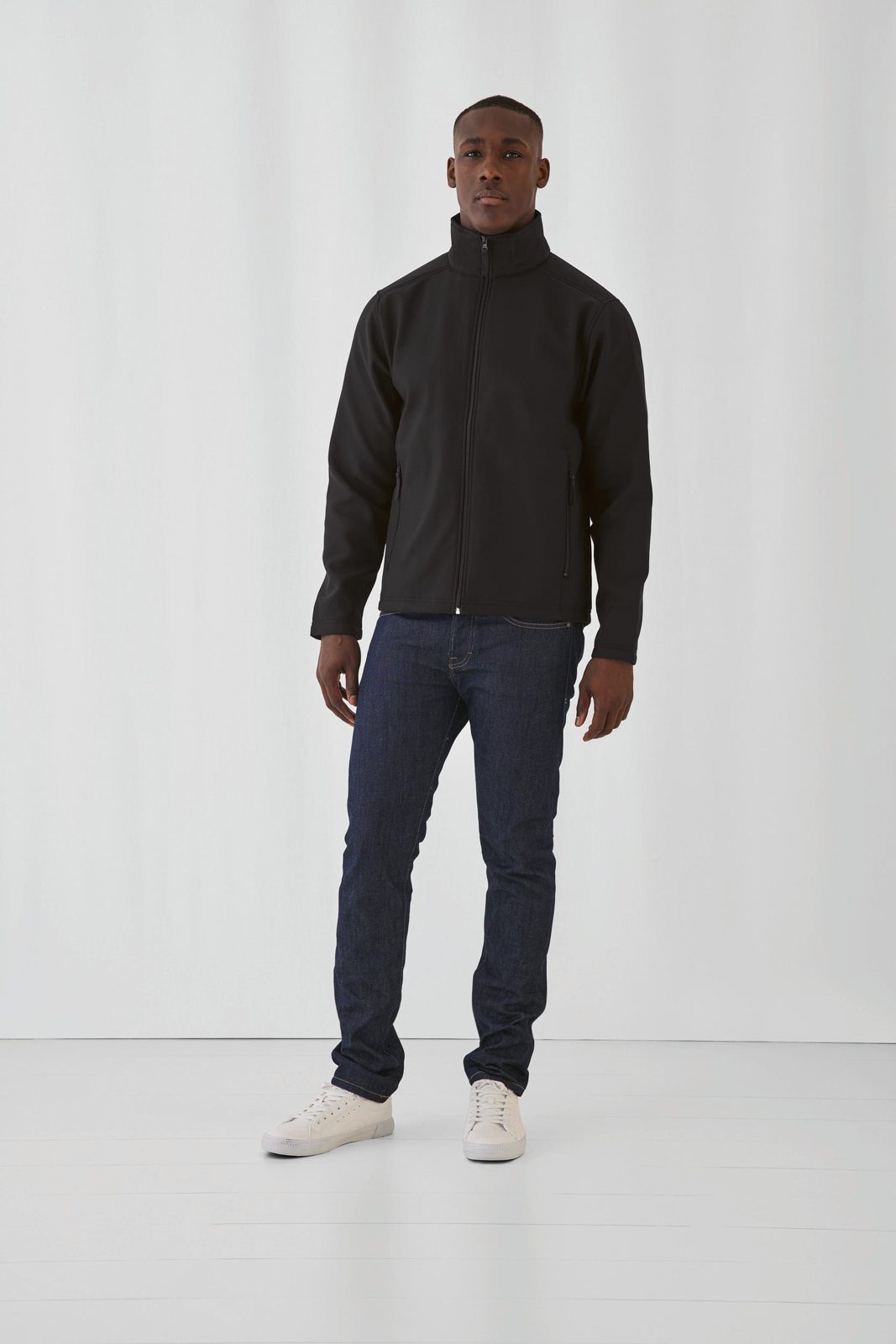 Man in een zwarte softshell jas met rits, spijkerbroek en witte sneakers
