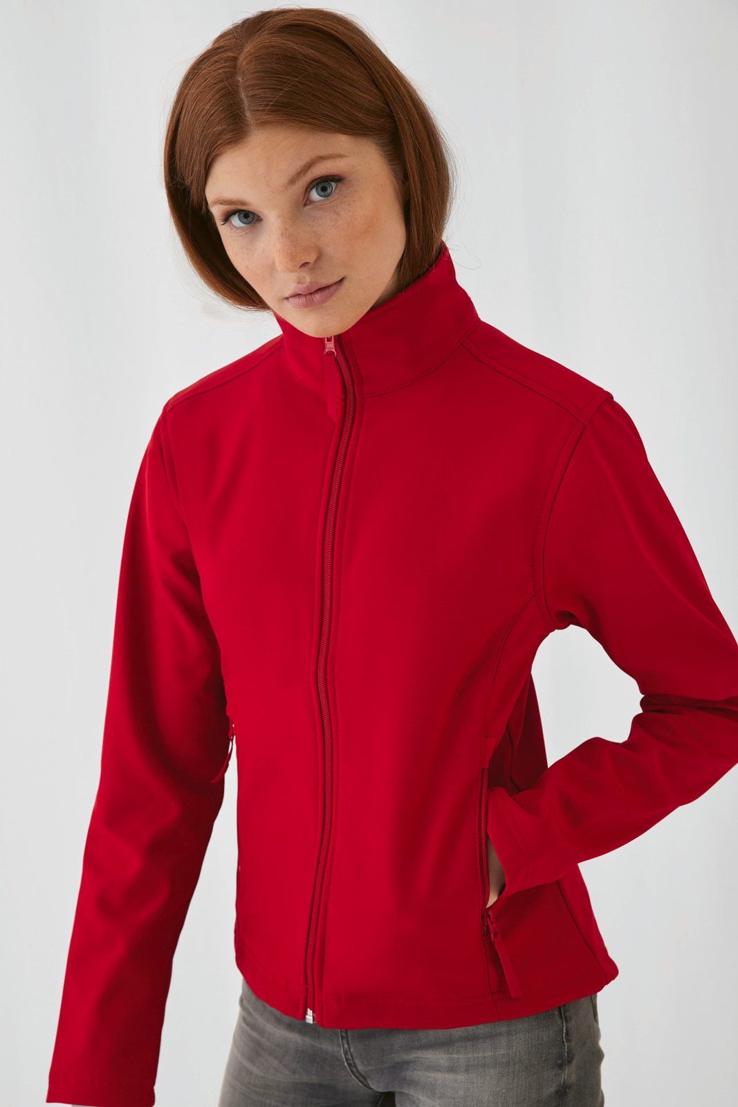 Vrouw met rood haar in een rode softshell jas met opstaande kraag, hand in de zak