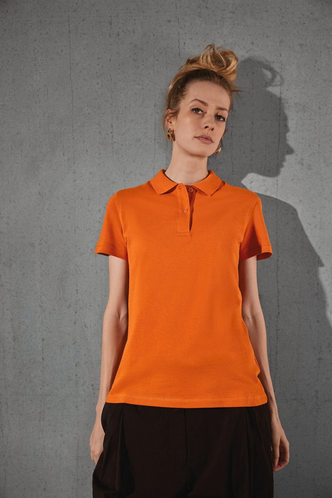 Vrouw met opgestoken haar in een oranje damespolo tegen een betonnen muur