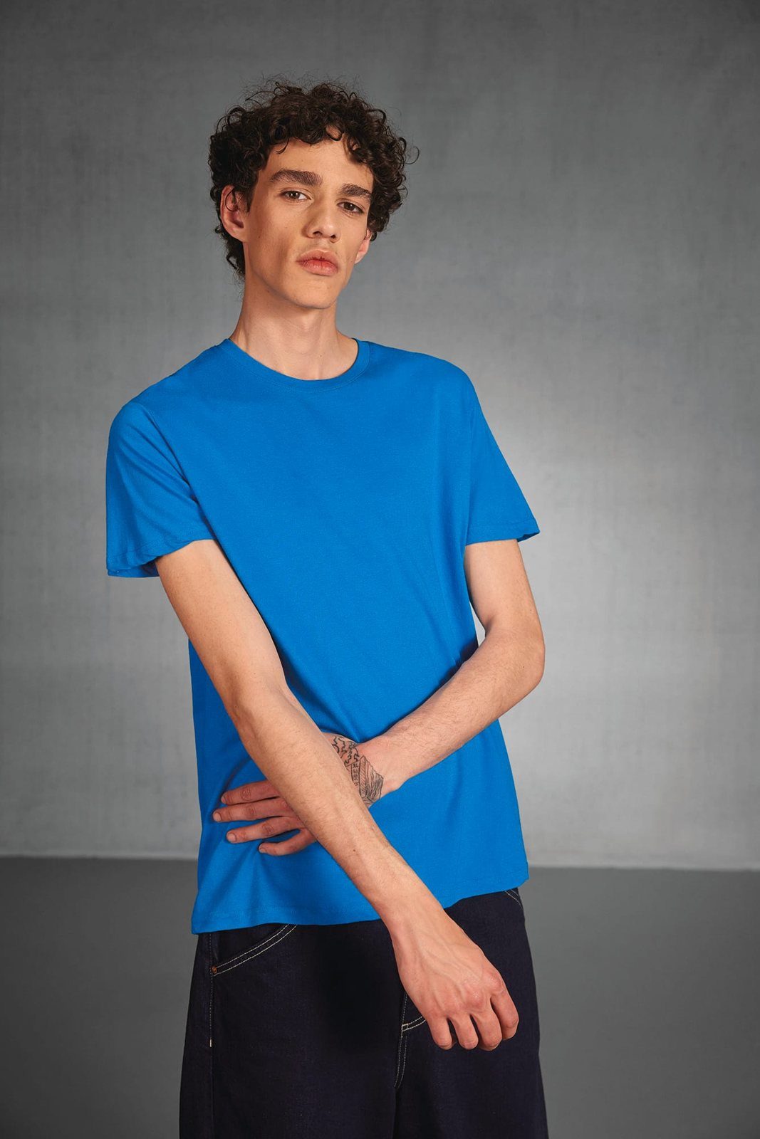 Jongeman met krullen in een felblauw T-shirt, arm voor zijn lichaam, grijze studiomuur
