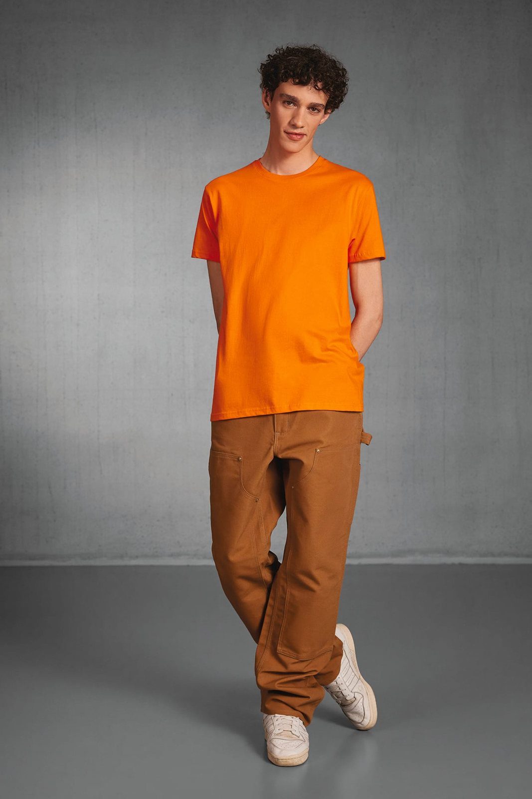Jongeman in een oranje T-shirt, bruine werkbroek en witte sneakers