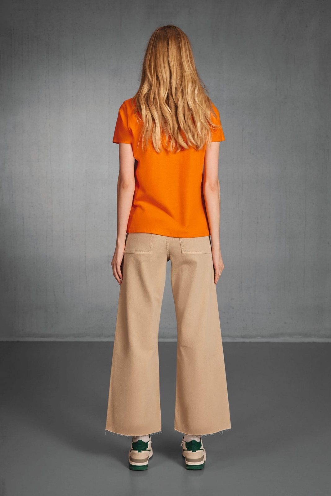 Achterkant van een vrouw met lang blond haar in een oranje T-shirt en wijde beige jeans