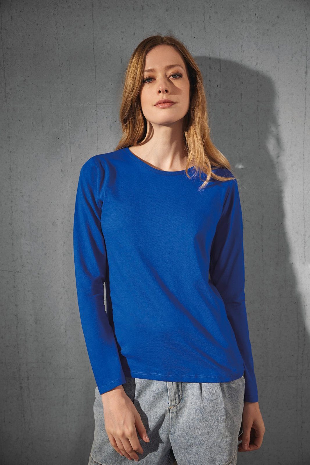Vrouw met lang haar in een kobaltblauw shirt met lange mouwen tegen een grijze muur