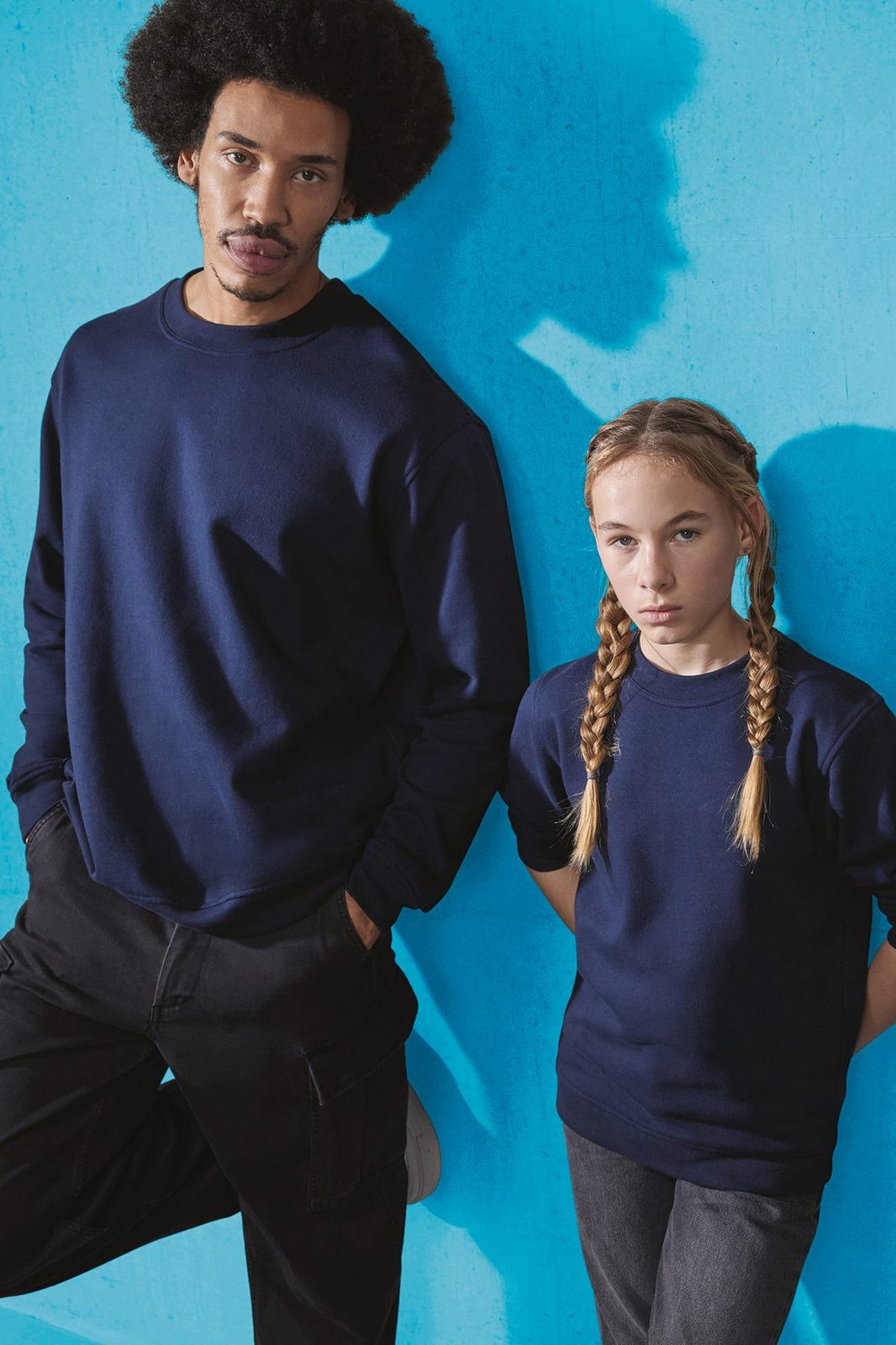 Man met afro en een meisje met vlechten in donkerblauwe sweaters tegen een blauwe muur
