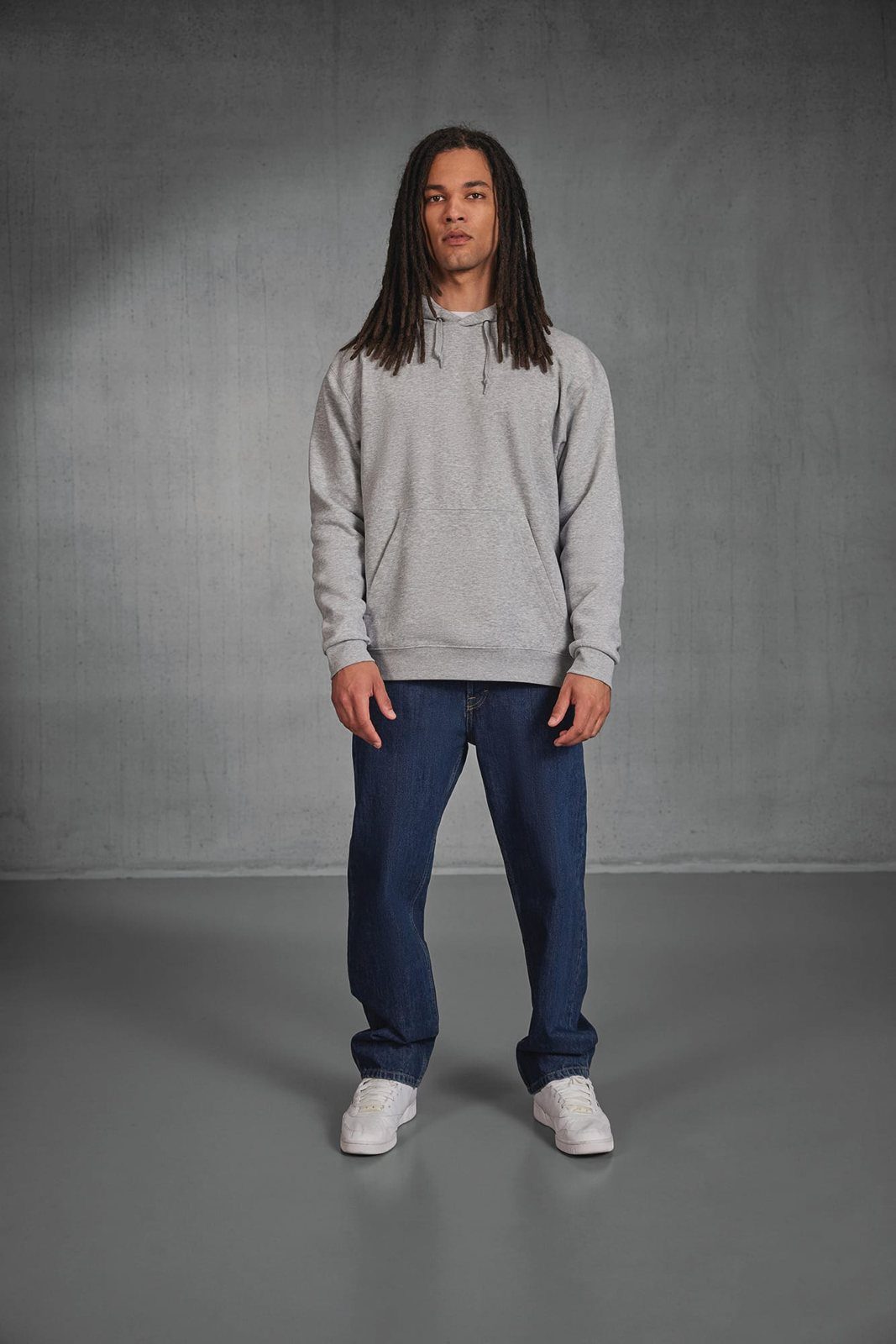 Man met lange dreadlocks in een grijze hoodie en een spijkerbroek tegen een betonnen muur