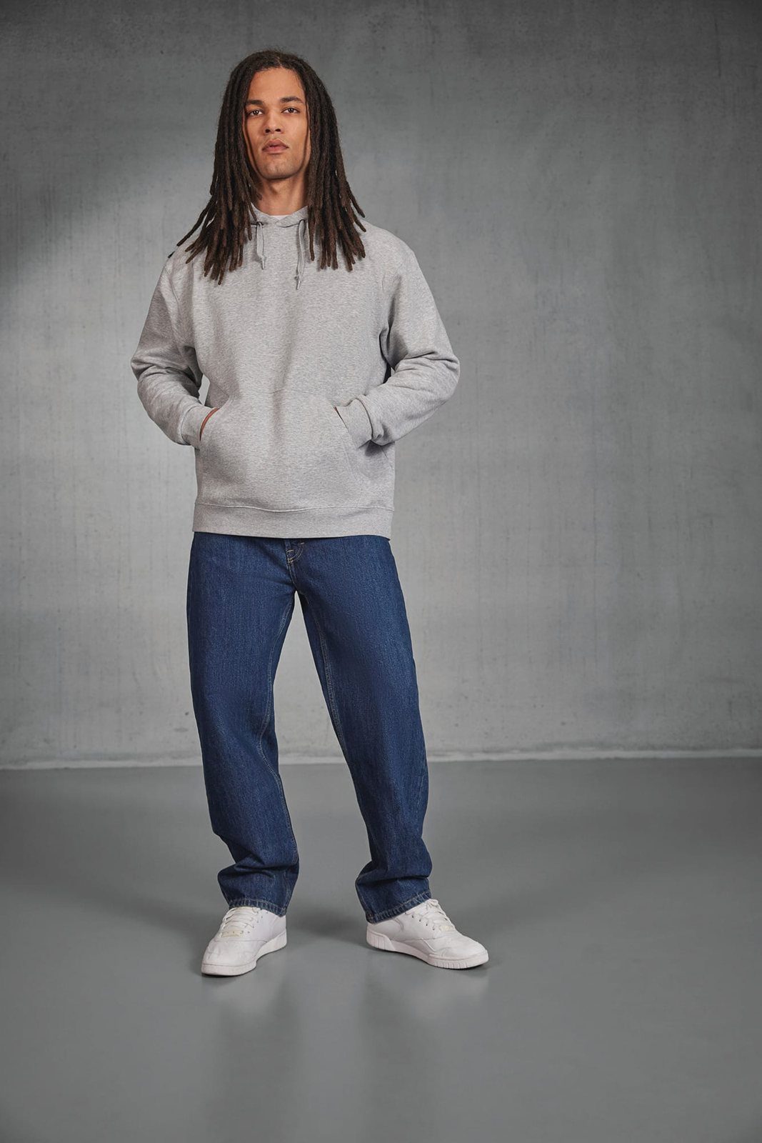 Man met lange dreadlocks in een lichtgrijze hoodie en spijkerbroek, handen in de buidelzak, in een betonnen studio