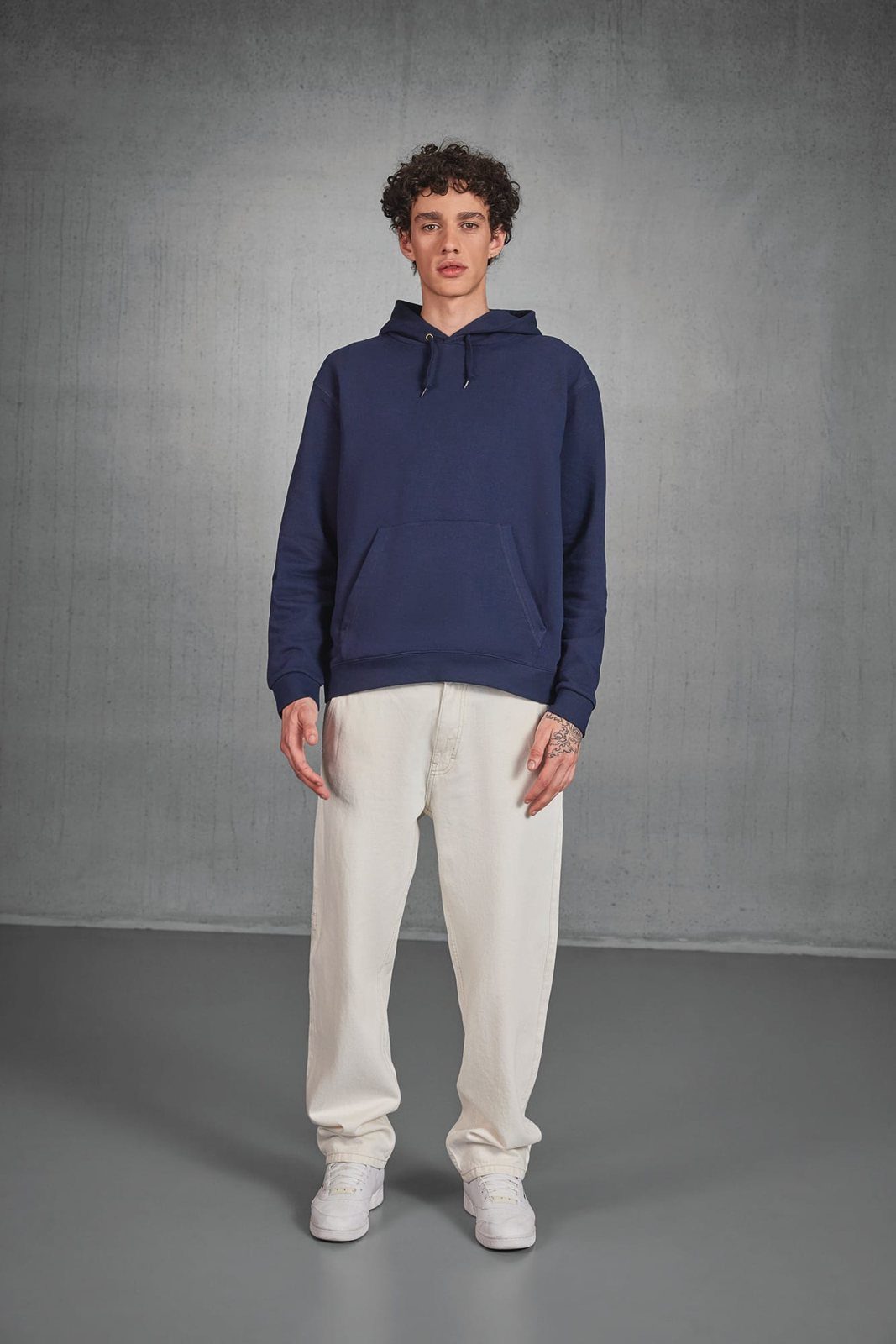 Jongeman met krullen in een donkerblauwe hoodie en een ruime crèmekleurige broek