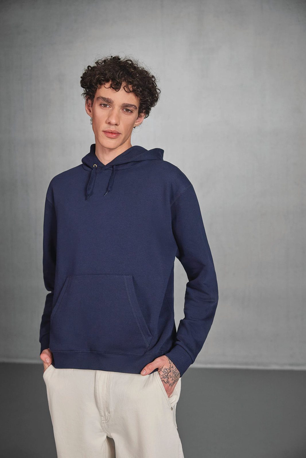 Jongeman met krullen in een donkerblauwe hoodie, handen in de zakken van een crèmekleurige broek