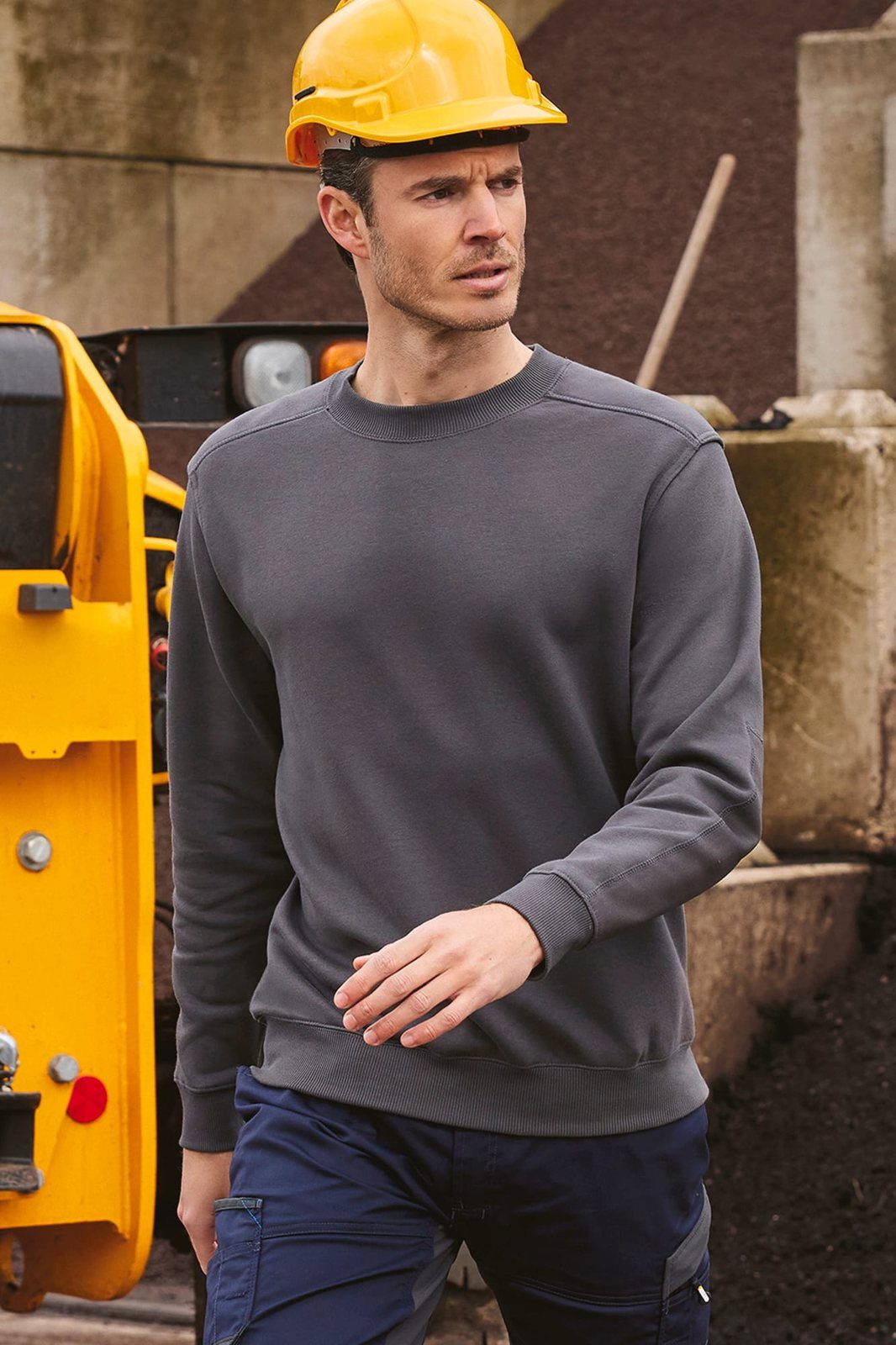 Man met gele bouwhelm in een grijze sweater met ronde hals naast een gele shovel