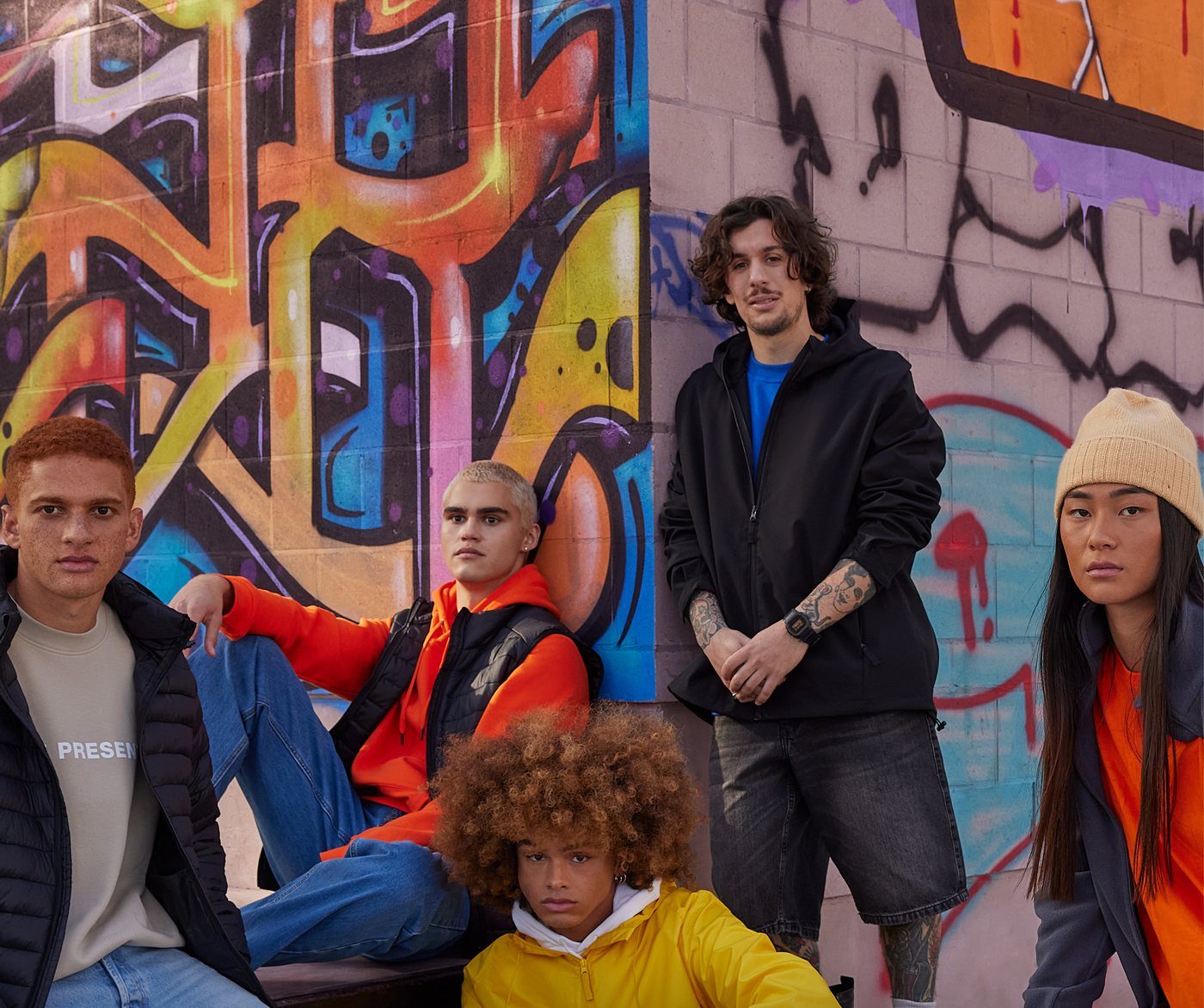 Vijf jongeren in jassen, hoodies en bodywarmers voor een muur vol graffiti