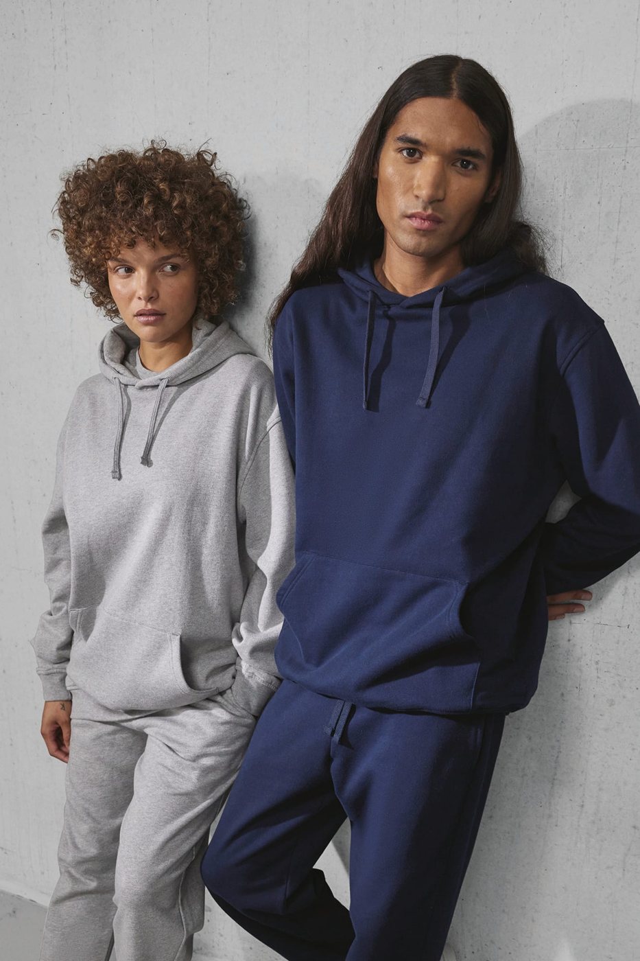 Vrouw in een grijze en man in een navy hoodie tegen een betonnen muur
