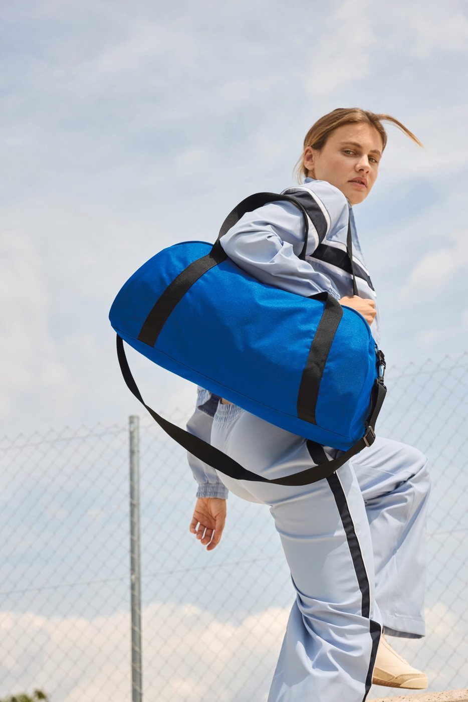 Vrouw in een lichtblauw trainingspak met een blauwe sporttas buiten