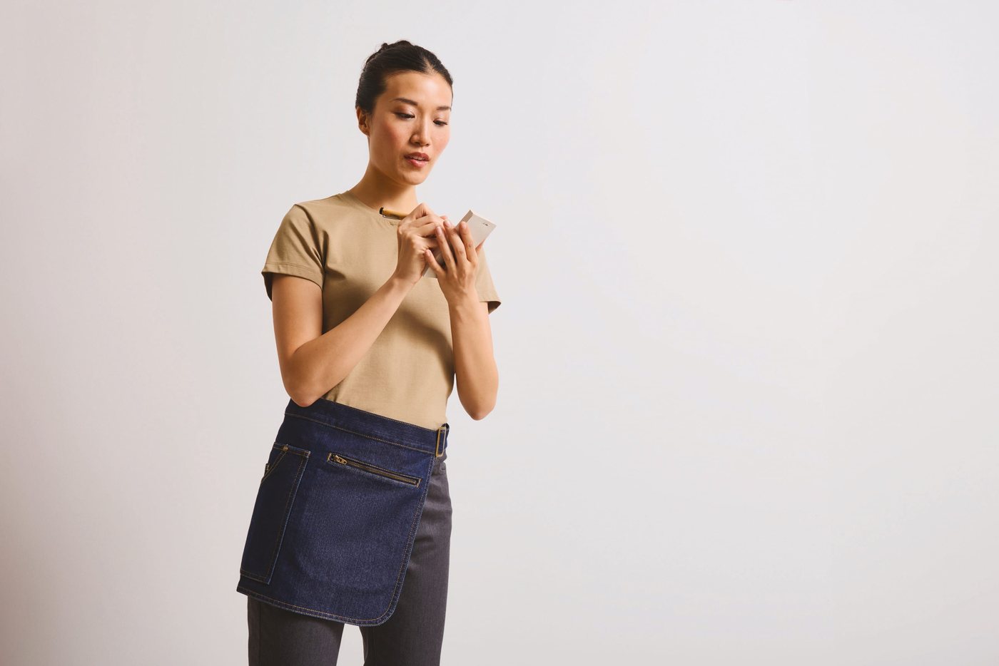 Vrouw in een beige T-shirt met een denim heupschort, kijkend op haar telefoon