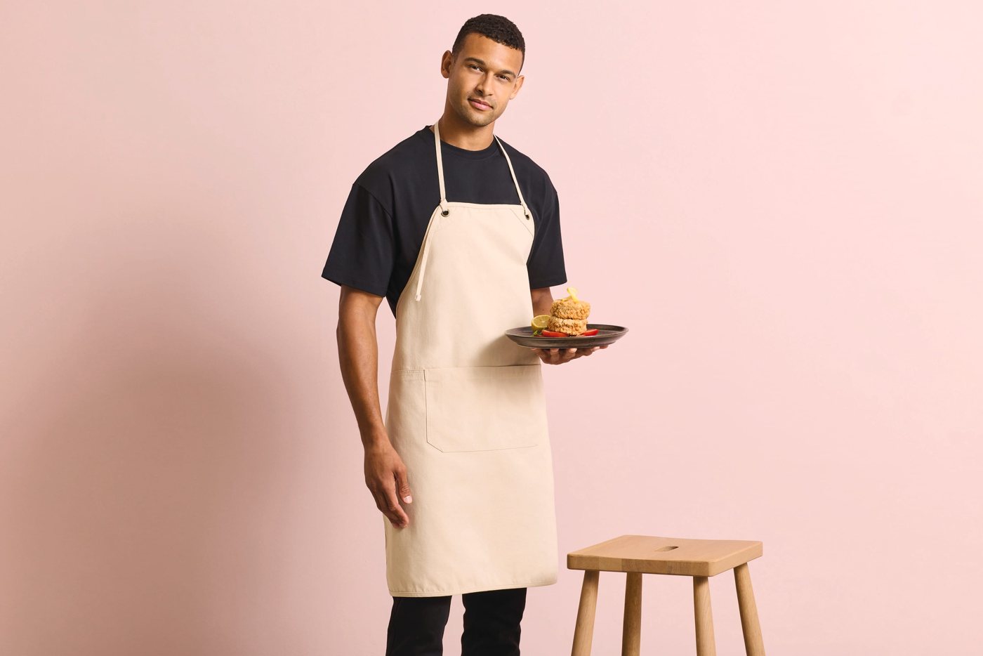 Man in een crèmekleurig canvas schort over een zwart T-shirt, met een bord eten, tegen een roze wand