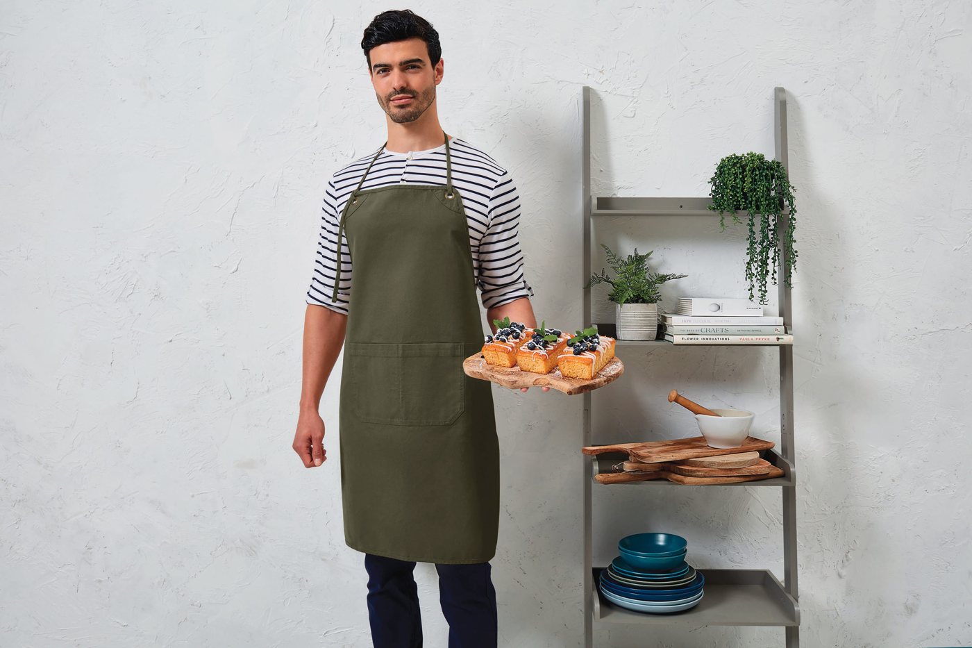 Man in een olijfgroen canvas schort over een gestreept shirt, met een plank gebak