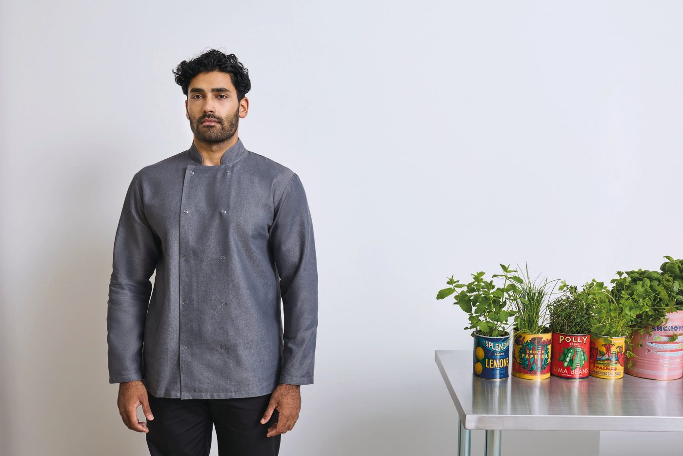 Man met krullen in een grijsblauwe denim koksjas, met kruidenpotten op de achtergrond